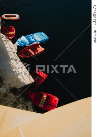 Several small colorful boats are floating near concrete dock, casting shadows on dark water below, creating tranquil and picturesque scene. Colorful boats floating near concrete dock 132605275