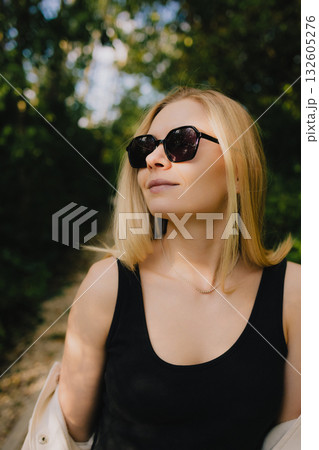 Young blonde woman with sunglasses enjoying summer day in park, wearing black tank top and white overshirt, looking up at sunlight filtering through trees. Portrait of blonde woman during summer day 132605276
