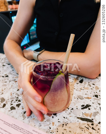 Woman is holding colorful cocktail with ice cubes, pear slice, and paper straw at modern bar, enjoying refreshing drink in stylish setting. Vibrant cocktail with pear garnish, ice, paper straw 132605286