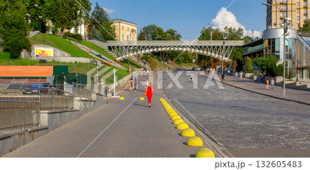 This image shows a pedestrian area, possibly a riverbank or waterside walkway, with a number of people walking or standing around. In the background, there is a bridge connecting two higher levels of 132605483