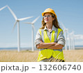A young woman in a yellow vest and hard hat stands proudly in a field with wind turbines in the background. 132606740