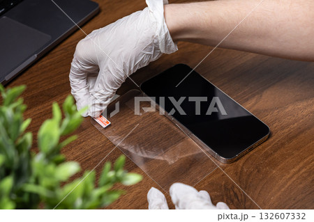 Hand in white glove peeling the backing from a transparent screen protector for precise application on a smartphone, ensuring gadget protection Hand in white glove peeling the backing from a transparent screen protector for precise application on a smartphone, ensuring gadget protection 132607332