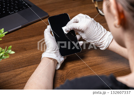Person in protective white gloves carefully cleans modern smartphone screen on wooden desk, highlighting essential digital device hygiene and health protection 132607337