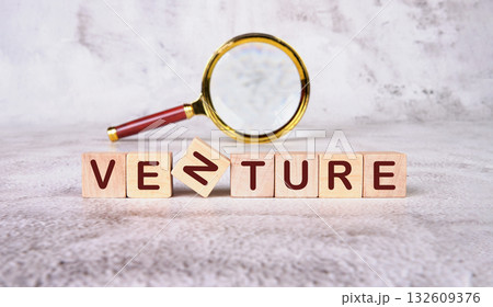 Wooden letter blocks spell out the word venture, with one slightly tilted. A gold and red magnifying glass hovers nearby, signifying a detailed examination of a new business endeavor 132609376