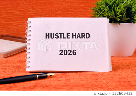 A notebook displays Hustle Hard 2026 on a desk. The setting includes a pot plant, a pen, and other office supplies on an orange surface A notebook displays Hustle Hard 2026 on a desk. The setting includes a pot plant, a pen, and other office supplies on an orange surface 132609412