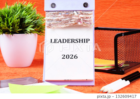 A desk calendar displays LEADERSHIP and 2026. Various desk supplies, a potted plant, and a black wire mesh organizer are arranged on an orange surface 132609417