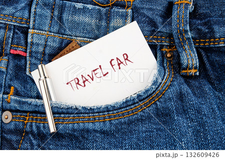 A small white note with the words TRAVEL FAR is tucked into the pocket of a pair of blue jeans. A silver pen rests against the pocket, suggesting readiness for exploration 132609426
