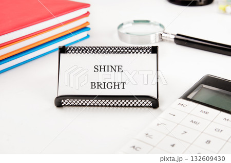 On a white desk, a black holder displays a card with the words SHINE BRIGHT. Surrounding it are colorful notebooks, a magnifying glass, and a white calculator in a bright, clean workspace On a white desk, a black holder displays a card with the words SHINE BRIGHT. Surrounding it are colorful notebooks, a magnifying glass, and a white calculator in a bright, clean workspace 132609430