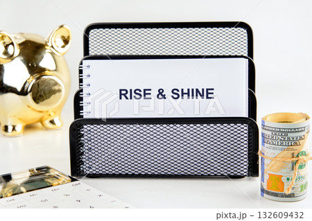 A notepad with the words RISE and SHINE sits within a black wire rack, displayed alongside a golden Piggy Bank, a roll of money, and a calculator A notepad with the words RISE and SHINE sits within a black wire rack, displayed alongside a golden Piggy Bank, a roll of money, and a calculator 132609432