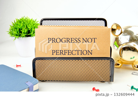 A kraft paper card rests in a black wire holder. Inscribed are the words PROGRESS NOT PERFECTION. This sits amongst a blue journal, pushpins, a plant, and a gold piggy bank A kraft paper card rests in a black wire holder. Inscribed are the words PROGRESS NOT PERFECTION. This sits amongst a blue journal, pushpins, a plant, and a gold piggy bank 132609444