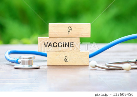 Wooden blocks spell out Vaccine with supporting hand gesture symbols. A Stethoscope lies nearby on a surface with a green, blurred background. Medicine and good health are conveyed 132609456