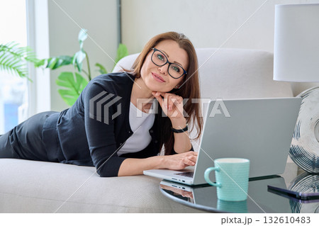 Portrait of middle aged woman with laptop lying on sofa at home Portrait of middle aged woman with laptop lying on sofa at home 132610483