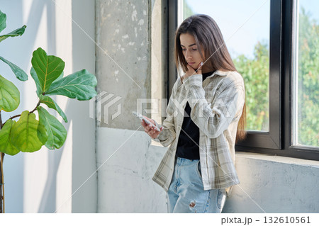 Serious teenage female standing at home reading texting on smartphone Serious teenage female standing at home reading texting on smartphone 132610561