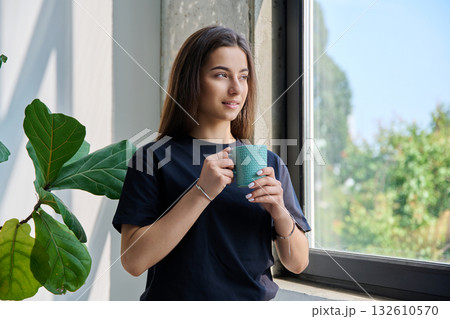 Portrait of smiling relaxed teenage girl posing with cup near window Portrait of smiling relaxed teenage girl posing with cup near window 132610570