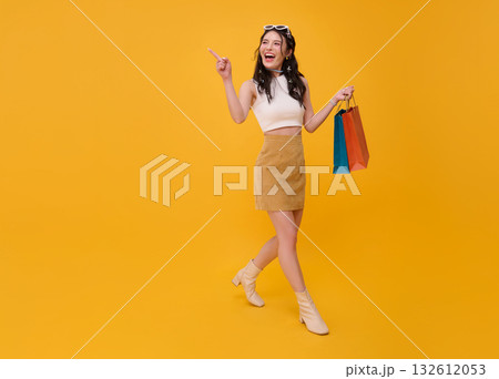 Happy young Asian woman shopper walking carrying shopping bags pointing finger up isolated on yellow copy space background. 132612053