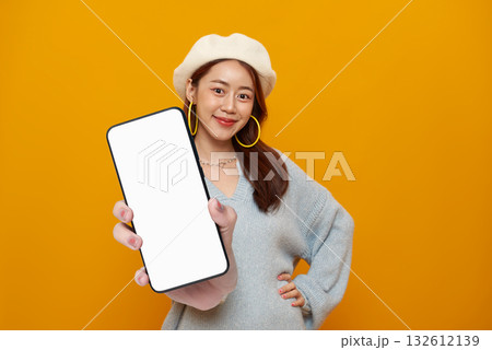 Smartphone mockup blank screen in hand cheerful Asian woman showcasing mobile app on vibrant yellow studio background for advertising and copy space 132612139