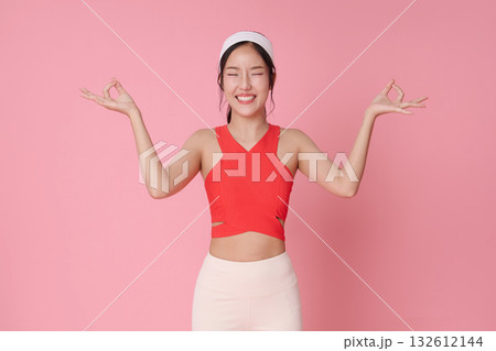 Fitness woman meditation pose with OK mudra on pink background smiling in red sports top for wellness yoga balance and healthy lifestyle advertising 132612144