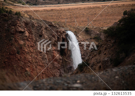 Waterfall pours over Rocky Cliff, surrounded by Rugged, arid landscape. Contrasting beauty of the flowing water against the dry, earthy tones of the canyon, nature's power and serenity 132613535