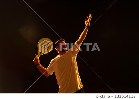 Man serves during a padel match in a dimly lit indoor court at night 132614118
