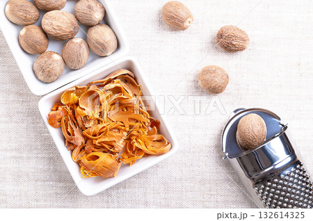 Dried mace, and a bunch of nutmegs, in white bowls, on linen. Spice made from reddish seed coverings of nutmeg seeds, with similar flavor to nutmeg, but more delicate, used for cooking and pickling. Dried mace, and a bunch of nutmegs, in white bowls, on linen. Spice made from reddish seed coverings of nutmeg seeds, with similar flavor to nutmeg, but more delicate, used for cooking and pickling. 132614325