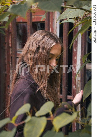 Teen girl with wavy hair stands near rusty metal bars, surrounded by green leaves, in soft natural light Teen girl with wavy hair stands near rusty metal bars, surrounded by green leaves, in soft natural light 132616931