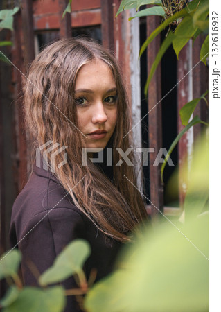 Teen girl with wavy hair stands near rusty metal bars, surrounded by green leaves, in soft natural light Teen girl with wavy hair stands near rusty metal bars, surrounded by green leaves, in soft natural light 132616932