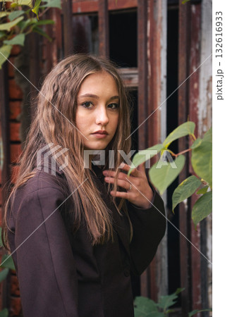Teen girl with wavy hair stands near rusty metal bars, surrounded by green leaves, in soft natural light 132616933