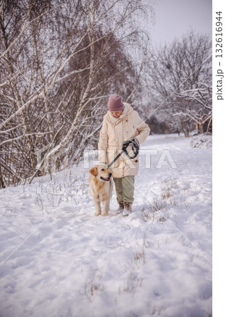 Elderly woman walking her Golden Retriever on a snowy countryside path. The woman gently looks at her dog while holding the leash, surrounded by snow-covered trees and soft winter sunlight. Elderly woman walking her Golden Retriever on a snowy countryside path. The woman gently looks at her dog while holding the leash, surrounded by snow-covered trees and soft winter sunlight. 132616944