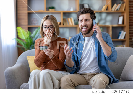 Confused woman holding her credit card and shocked by a fraudulent transaction while her angry husband is making a phone call, shouting about financial problems Confused woman holding her credit card and shocked by a fraudulent transaction while her angry husband is making a phone call, shouting about financial problems 132617757