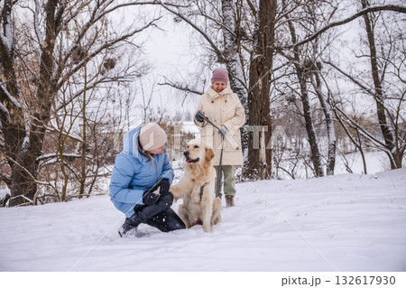 Elderly woman and her adult daughter spending a winter day outdoors with their Golden Retriever. The daughter kneels beside the dog, petting it affectionately, while the mother holds the leash amid a Elderly woman and her adult daughter spending a winter day outdoors with their Golden Retriever. The daughter kneels beside the dog, petting it affectionately, while the mother holds the leash amid a 132617930