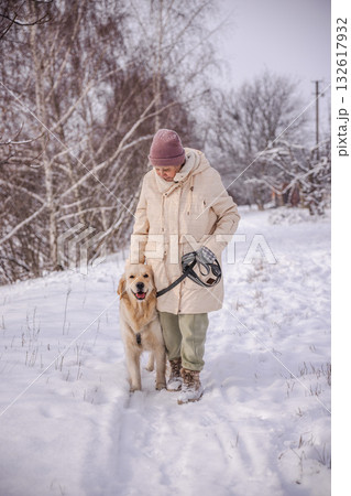 Elderly woman walking her Golden Retriever on a snowy path in the countryside. The woman looks down at her loyal dog with affection, enjoying a quiet winter day surrounded by snow and bare trees. Elderly woman walking her Golden Retriever on a snowy path in the countryside. The woman looks down at her loyal dog with affection, enjoying a quiet winter day surrounded by snow and bare trees. 132617932