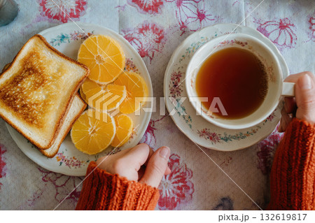 Toasted bread, sliced orange, and hot tea on floral tablecloth create cozy breakfast scene with warm, inviting colors and relaxed morning atmosphere 132619817