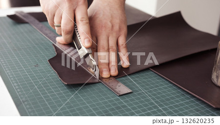 Leather craftsman cutting brown leather sheet with utility knife and metal ruler 132620235