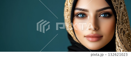 Beautiful woman with striking blue eyes, wearing a black hijab with gold embroidery, poses against a teal backdrop 132621023