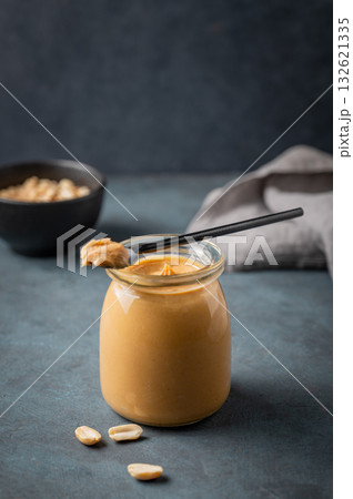 A jar of creamy peanut butter on a dark background with a scattering of roasted nuts, spoon 132621335