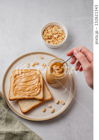 A hand holds a spoon with creamy peanut butter in a jar on a plate with toast and roasted nuts A hand holds a spoon with creamy peanut butter in a jar on a plate with toast and roasted nuts 132621343
