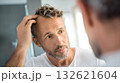 Middle aged man with graying hair and beard is touching his hair and looking in the mirror, examining his hairline and scalp for hair loss or thinning 132621604