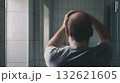 Upset man examining his hair loss in the bathroom mirror, touching his scalp and grappling with the challenges of alopecia and thinning hair during his morning routine 132621605