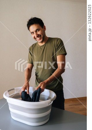 Vertical portrait of cheerful young man washing clothes by hand in plastic basin, wringing out water performing domestic task, smiling looking at camera. Concept of hygiene and cleanliness. 132622025