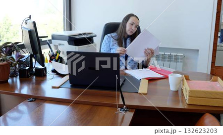 Focused businesswoman reviewing paperwork in a busy office, surrounded by equipment and supplies. Natural light creates a productive atmosphere for efficient work Focused businesswoman reviewing paperwork in a busy office, surrounded by equipment and supplies. Natural light creates a productive atmosphere for efficient work 132623203