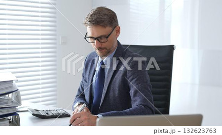 Mature entrepreneur in suit, tie and glasses is sitting at desk, writing notes and using calculator in modern, bright office. Audit and taxes in business 132623763