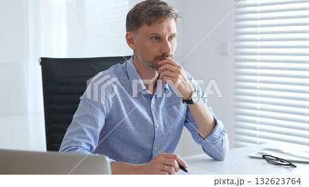 Businessman wearing blue sleek shirt is thinking and concentrating while working at the desk in a bright, modern office, demonstrating professionalism and efficiency. Business people concept 132623764