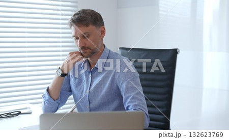 Businessman wearing blue sleek shirt is thinking and concentrating while working at the desk in a bright, modern office, demonstrating professionalism and efficiency. Business people concept 132623769