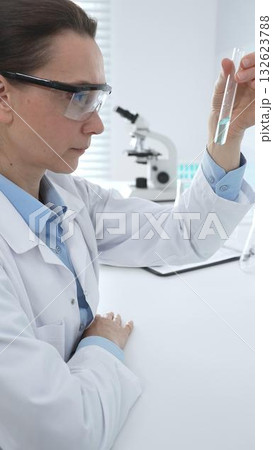 Female researcher examining clear liquid in test tube, sitting near microscopes and lab equipment within sterile scientific workspace. Medicine, healthcare and science concept Female researcher examining clear liquid in test tube, sitting near microscopes and lab equipment within sterile scientific workspace. Medicine, healthcare and science concept 132623788