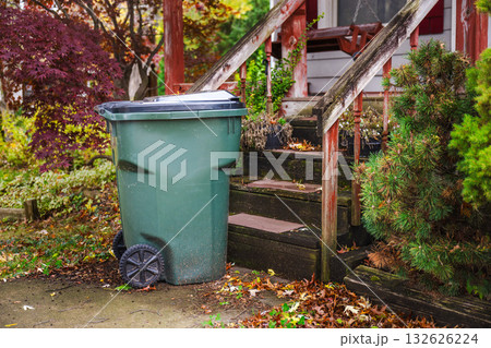 Large green garbage bin on wheels standing near the front steps of an old wooden porch surrounded by autumn foliage. Large green garbage bin on wheels standing near the front steps of an old wooden porch surrounded by autumn foliage. 132626224