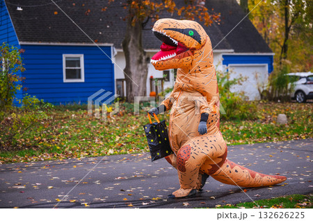 Person in inflatable dinosaur costume walking along suburban street with a Halloween candy bag. Person in inflatable dinosaur costume walking along suburban street with a Halloween candy bag. 132626225
