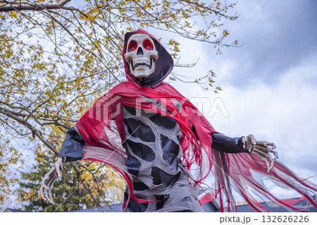 Creepy Halloween skeleton decoration with glowing red eyes and torn red cloak under cloudy autumn sky. Creepy Halloween skeleton decoration with glowing red eyes and torn red cloak under cloudy autumn sky. 132626226