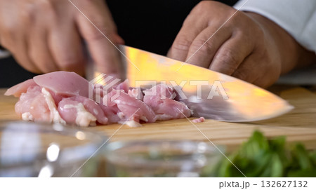 A chef using a kitchen knife slices uncooked chicken meat on a chopping board, the close up view of preparing food ingredients. A chef using a kitchen knife slices uncooked chicken meat on a chopping board, the close up view of preparing food ingredients. 132627132