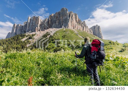 Hiker man with trekking poles, backpack and solar panel in green meadow with Bolshoy Tkhach mountain Hiker man with trekking poles, backpack and solar panel in green meadow with Bolshoy Tkhach mountain 132634430
