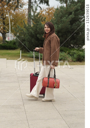 A stylish woman prepares to travel, holding luggage near a modern building. The image combines fashion, confidence, and the mood of departure. A stylish woman prepares to travel, holding luggage near a modern building. The image combines fashion, confidence, and the mood of departure. 132639018